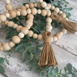 May include: A wooden bead garland with two brown jute tassels. The garland is made of natural wood beads and is strung on a natural twine. The garland is displayed on a whitewashed wood surface with green foliage.