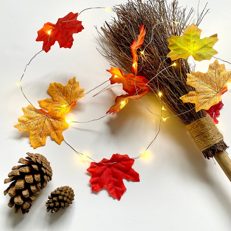 May include: A brown broom with a rustic handle and a string of fairy lights wrapped around it. The lights are strung with fall-colored leaves, including red, orange, and yellow.