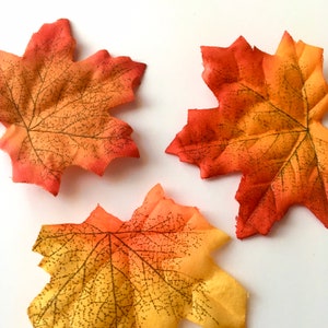May include: Three artificial fall leaves in shades of orange, yellow, and red. The leaves have a detailed vein pattern and are made of a fabric-like material.
