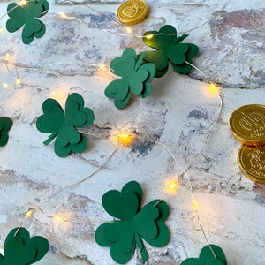 May include: Green paper shamrocks strung with white fairy lights on a whitewashed wood background. Gold coins are also strung on the lights.