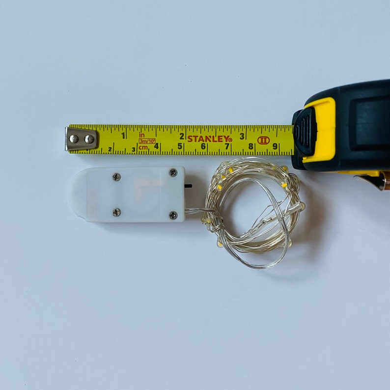 May include: A white battery-powered LED light string with a clear wire and warm white lights. The string is coiled up and next to a white battery pack with a yellow tape measure labeled "STANLEY" in the background.