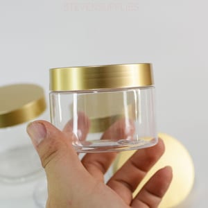 以下が含まれることがあります： 手で持たれた、金色の蓋が付いた透明なプラスチック製の瓶。瓶は円筒形で透明で、中身が見えます。蓋は光沢のある金色です。「STEVENSSUPPLIES」の文字が上部に表示されています。