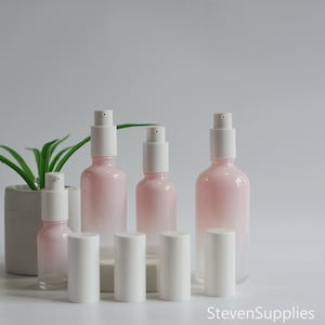 May include: Set of four pink glass bottles with white pump tops. The bottles are ombre, with a darker pink at the bottom and a lighter pink at the top. The bottles are arranged in a row, with the smallest bottle on the left and the largest bottle on the right.