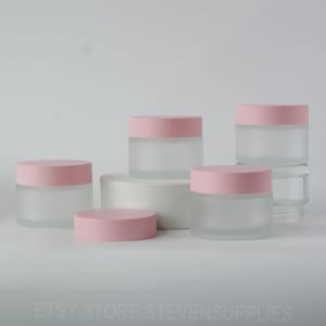 Op de afbeelding: Vijf frosted glazen potten met roze deksels. De potten zijn van verschillende grootte en zijn gerangschikt op een wit oppervlak.