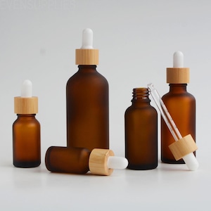 May include: A collection of amber glass dropper bottles with bamboo caps and white droppers. The bottles are in different sizes, some standing and some lying down. One bottle has a dropper inserted, and another has the dropper removed.