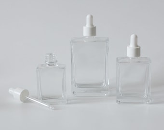 May include: Three clear glass bottles with white dropper tops. The bottles are different sizes and are arranged in a row on a white surface.