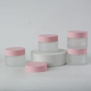 Op de afbeelding: Vijf frosted glazen potten met roze deksels. De potten zijn gerangschikt op een wit oppervlak. De potten zijn van verschillende grootte.