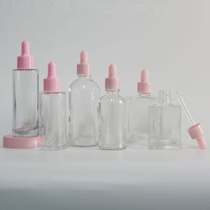 May include: Clear glass dropper bottles with pink tops and droppers. The bottles vary in shape and size, with a few round and square options. One bottle has a pink base. The bottles are arranged on a white surface.