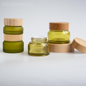 Puede incluir: Varios frascos de vidrio verde oliva con tapas de bambú. Un frasco está abierto, revelando una crema blanca. Los frascos están apilados y dispuestos sobre una superficie blanca. El texto "STEVENSUPPLIES" está en la esquina superior izquierda.