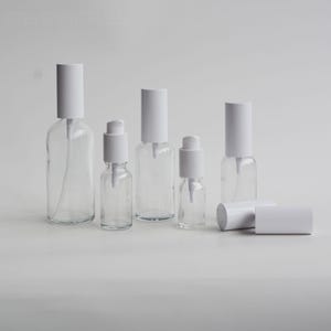 Op de afbeelding: Transparante glazen flessen met witte pompkappen en witte doppen. De flessen variëren in grootte, sommige staan rechtop en andere liggen. De achtergrond is effen wit.
