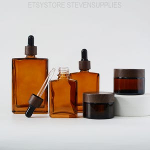 Puede incluir: Cuatro botellas de vidrio ámbar con tapas de madera y goteros, y un frasco de vidrio marrón con tapa de madera. Las botellas y el frasco están dispuestos sobre una superficie blanca.