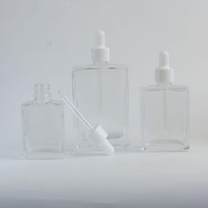 May include: Clear glass dropper bottles with white tops in various sizes. One bottle is open, with a white dropper resting on the surface. The bottles are rectangular with rounded corners, suitable for essential oils or serums.