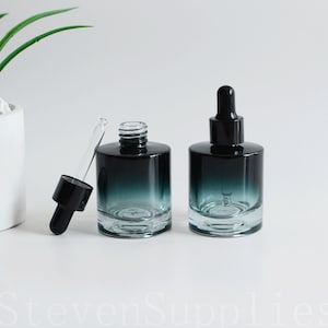 Puede incluir: Dos frascos cuentagotas de vidrio con un diseño degradado en verde azulado y negro. Un frasco tiene un cuentagotas negro insertado, el otro está abierto. Los frascos son cilíndricos con una base de vidrio transparente.