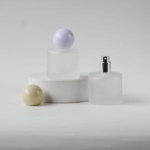 May include: Two frosted glass perfume bottles with white and silver accents. One bottle has a white spherical cap, the other has a silver spray top. A beige sphere and a white cylindrical pedestal are also present.
