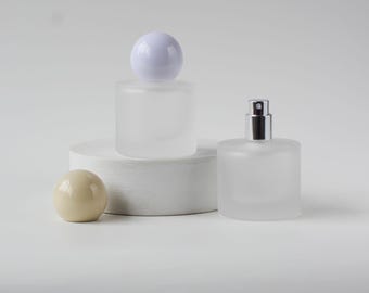 May include: Two frosted glass perfume bottles with white and silver accents. One bottle has a white spherical cap, the other has a silver spray top. A beige sphere and a white cylindrical pedestal are also present.