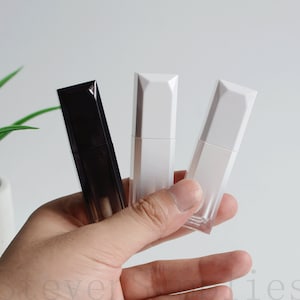 Puede incluir: Tres tubos de lápiz labial en una mano. Uno es negro, los otros dos son blancos y transparentes. Los tubos tienen un diseño geométrico. El fondo es blanco.