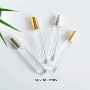Puede incluir: Cuatro frascos de perfume de vidrio transparente con tapas plateadas y doradas. Los frascos están dispuestos en forma de V sobre un fondo blanco. El texto "STEVENSUPPLIES" está impreso debajo de los frascos.