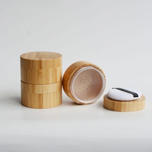 May include: Three bamboo powder containers. Two are stacked, one is open, revealing a sifter and powder. A white powder puff with a black band sits on a bamboo lid. The containers are cylindrical and made of light brown bamboo.