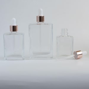 May include: Three clear glass dropper bottles with rose gold collars and white droppers. The bottles are rectangular and vary in size. One bottle has its dropper removed and lying beside it.