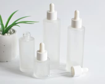 May include: Four frosted glass bottles with white dropper tops. The bottles are various sizes and are arranged on a white surface. The bottles are empty and ready to be filled with essential oils, serums, or other liquids.