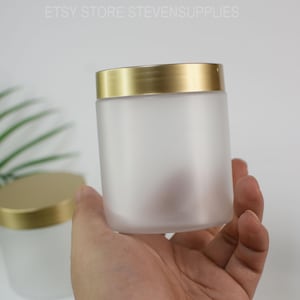 Op de afbeelding: Een pot van matglas met een goudkleurige deksel wordt in een hand gehouden. De pot is cilindrisch en ondoorzichtig, met een gladde, matte afwerking. De deksel heeft een glanzende goudkleur. De tekst "ETSY STORE STEVENSSUPPLIES" is bovenaan zichtbaar.