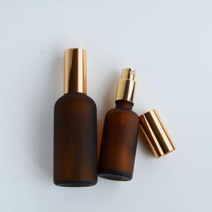Peut inclure: Deux flacons pulvérisateurs en verre brun givré avec des bouchons dorés. Un flacon a son bouchon, et l'autre a son bouchon enlevé, à côté du flacon. Les flacons sont sur fond blanc.