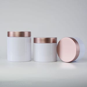 May include: Three white jars with rose gold lids. The jars are different sizes and are arranged in a row from largest to smallest.