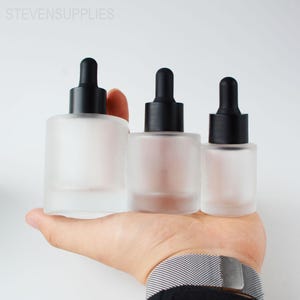 Op de afbeelding: Drie glazen druppelflessen met zwarte doppen worden in een hand gehouden. De flessen zijn cilindrisch en verschillen in grootte. De tekst "STEVENSSUPPLIES" is zichtbaar bovenaan de afbeelding.
