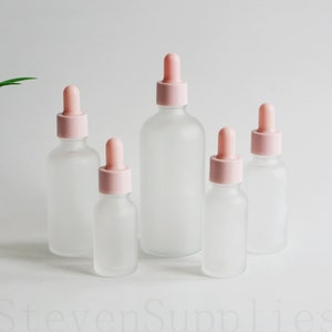 May include: Five frosted glass bottles with pink dropper tops. The bottles are different sizes and are arranged in a row from largest to smallest.