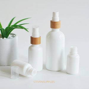 May include: Set of three white glass bottles with bamboo lids. The bottles have a frosted finish and are ideal for essential oils, serums, or other liquids. The bottles are arranged in a row on a white surface.