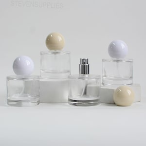 Puede incluir: Botellas de perfume de vidrio transparente con tapones esféricos en blanco y beige. Una botella tiene un atomizador plateado. Las botellas son cilíndricas y están sobre pedestales blancos. El texto "STEVENSUPPLIES" es visible.