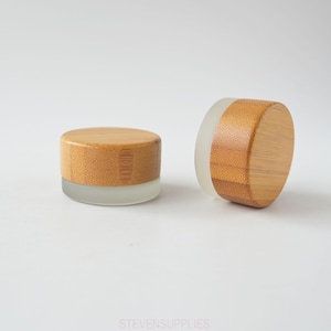 May include: Two small, round containers. Each has a bamboo lid and a frosted glass base. One container is upright, the other is tilted. The bamboo lids have a natural wood grain, and the glass bases are translucent.