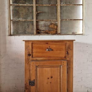 May include: A light brown wooden cabinet featuring a single top drawer with a metal pull handle and a lower cabinet door with a dark metal latch. The cabinet is constructed from natural wood with visible grain patterns and knots throughout the surface.