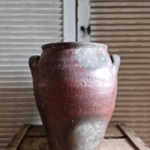 以下が含まれることがあります： 赤茶色の釉薬が施された、素朴でアンティーク風の陶器の壺。 2つの小さな取っ手があり、手作りのような、わずかに不規則な形状をしています。 壺の下部は、より明るく斑点のある外観をしています。