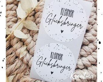 Può includere: Due adesivi rotondi bianchi con macchie nere e il testo "KLEINER Glücksbringer" in scrittura corsiva nera. Un piccolo cuore nero si trova sotto il testo su ogni adesivo.