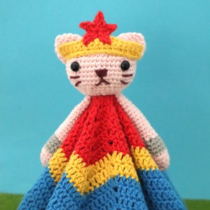 May include: A crocheted cat toy with a red, yellow, and blue blanket. The cat is wearing a yellow crown with a red star on top.
