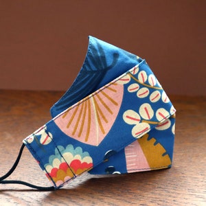 Op de afbeelding: Een blauw stoffen mondkapje met een bloemenpatroon in roze, wit en goud. Het masker heeft een zwart elastiek.