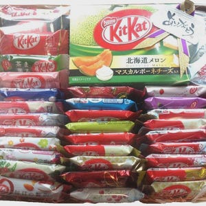 Può includere: Un cesto regalo pieno di varie barrette di cioccolato Kit Kat. Le barrette sono in una varietà di colori, tra cui rosso, verde, rosa e blu. La confezione include testo giapponese e immagini di frutta. È visibile una grande scatola di Kit Kat al gusto melone.