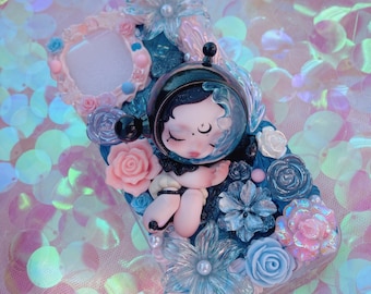 Puede incluir: Funda de teléfono decorativa con una figura de astronauta durmiente en un casco transparente, rodeada de decoraciones florales azules y rosas. La funda tiene un marco rosa claro y está adornada con varias flores.