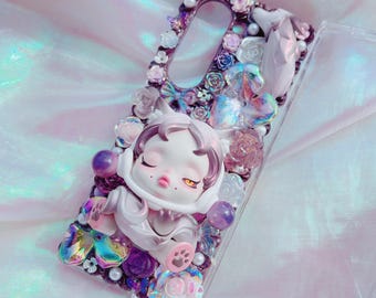 Puede incluir: Funda de teléfono transparente adornada con una figura 3D de una muñeca durmiente con orejas de gato, rodeada de adornos florales morados, rosas y blancos, perlas y detalles iridiscentes. La funda tiene un marco decorativo.