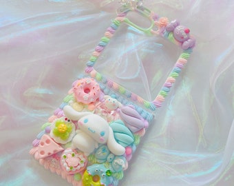 Pode incluir: Uma capa transparente para telefone decorada com glacê pastel, granulado e um cão de desenho animado branco. A capa apresenta uma variedade de doces e guloseimas coloridas, incluindo donuts, uma fatia de bolo e uma fruta verde e amarela.