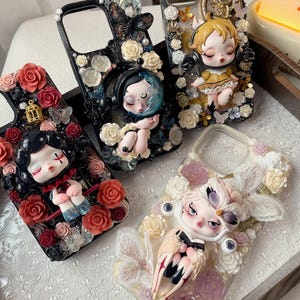 Puede incluir: Cuatro fundas de teléfono decorativas con figuras de muñecas detalladas y diseños florales. Las fundas presentan rosas, mariposas y otros adornos en varios colores, incluyendo negro, rojo y blanco. Los diseños son intrincados y artísticos.