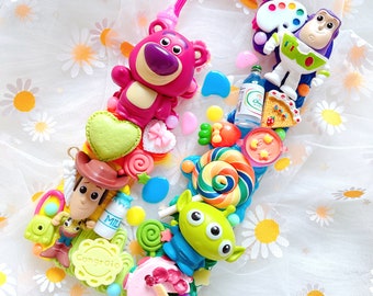 May include: A decorative phone case featuring various Toy Story characters and candy-themed embellishments. The case includes a pink Lotso bear, a green alien, and a Buzz Lightyear figure, along with colorful candies and a heart-shaped design.