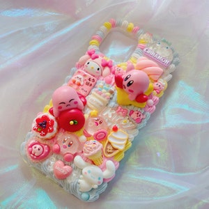 May include: A pink and white phone case decorated with 3D characters and food items. The case features characters from the video game Kirby and the anime Hello Kitty. The case also features a variety of food items, including ice cream, sweets, and cake.