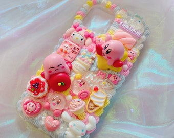 以下が含まれることがあります： ピンクと白の携帯電話ケースで、3Dのキャラクターと食べ物で飾られています。ケースには、ゲーム「カービィ」とアニメ「ハローキティ」のキャラクターが描かれています。ケースには、アイスクリーム、キャンディー、ケーキなど、さまざまな食べ物も描かれています。