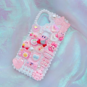 Puede incluir: Una funda de teléfono rosa y blanca decorada con varios abalorios 3D. La funda presenta un borde de encaje blanco y está adornada con corazones rosas, perlas y personajes de dibujos animados. El diseño incluye un lazo, fresas y un castillo en miniatura.
