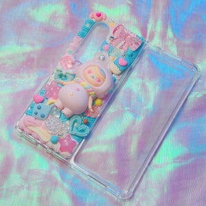 May include: A clear phone case with a colorful, 3D design featuring a pink and blue character with a candy cane and hearts. The design is made with resin and other embellishments.
