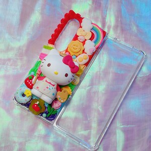 May include: A clear phone case decorated with a 3D design featuring Hello Kitty, a rainbow, a smiling sun, clouds, and a strawberry. The case has a red border and a variety of colorful elements.