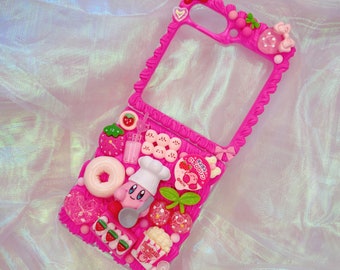Pode incluir: Uma capa de telefone rosa com a parte de trás transparente e um design rosa e branco. A capa é decorada com vários pingentes, incluindo um donut rosa e branco, um morango rosa, um chapéu de cozinheiro rosa e branco, um personagem rosa e branco com uma colher rosa, um balde de pipoca rosa e branco e um morango rosa e branco. A capa também tem um pingente de coração rosa e branco e um pingente de laço rosa e branco. A capa está sobre um fundo rosa e branco.