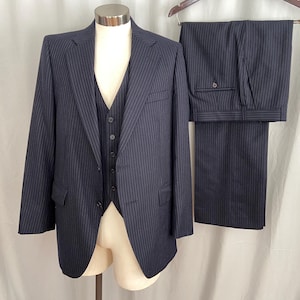May include: A navy blue, three-piece suit with a pinstripe pattern. The suit includes a blazer, vest, and trousers. The blazer is buttoned and displayed on a mannequin. The trousers are hanging on a wooden hanger.